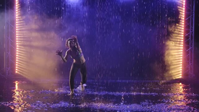 A silhouette of a sporty woman showing the technique of capoeira martial art in the rays of blue light under streams of rain. Slow motion.