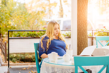 Charming serene pregnant woman in blue dress eating appetizing cake with cream in outdoor cafe or terrace of restaurant.