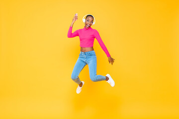 Fototapeta na wymiar Full length portrait of jumping happy energetic African American woman wearing headphones listining to music from smartphone isolated on colorful yellow background