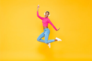 Fototapeta na wymiar Fun portrait of smiling energetic African American woman wearing headphones listining to music from smartphone and jumping isolated on colorful yellow background