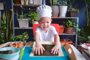 Cute happy smiling Asian little boy child wearing chef hat having fun preparing, cooking healthy Japanese food -  sushi roll at home, Fun activities for kindergarten kids concept