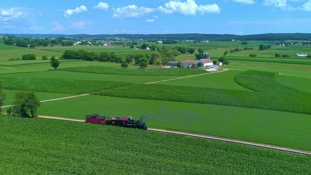 Aerial View of an Antique Steam Engine and Caboose Steaming and Puffing across Green Farmlands
