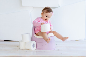 a little girl sits on a potty in pink clothes in a bright room and plays with toilet paper