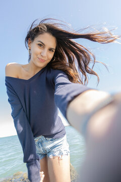 Vertical shot selfie of young brunette italian woman long hair dark blue sweater romantic hair in the wind happy, smiling and with the intense blue sky and with a flower in her right hand holding down