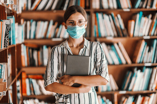 Beautiful intelligent freshman girl with face mask on standing in library and holding tablet in hands. remote studying during corona virus concept.