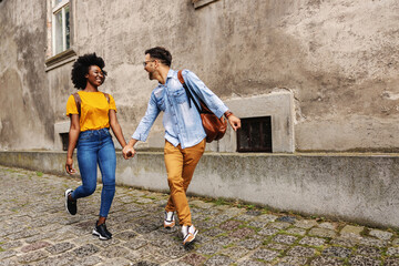 Young cheerful multicultural hipster couple walking in an old part of the town and holding hands.