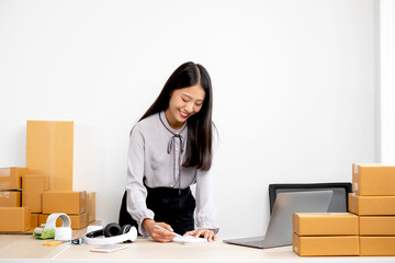 Young Asian woman business owner with many parcel boxes on the table happy online sales job, use your laptop, get an order from customers, take notes, and make arrangements for delivery by post