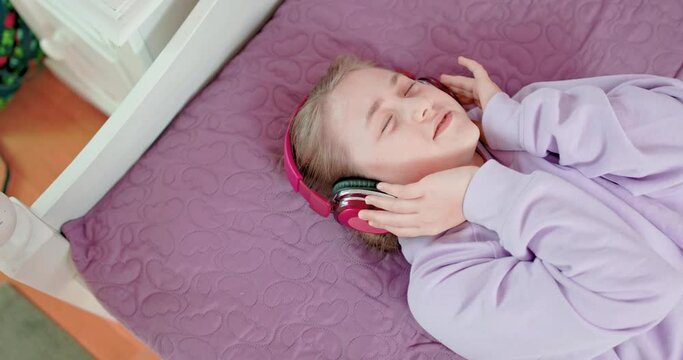 Teen girl is singing with closed eyes lying on her bed in bedroom wearing headphones and waving hands