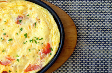Yellow omelet with tomato and bacon slices, sprinkled with herbs, in a round black skillet on a brown oval wooden stand