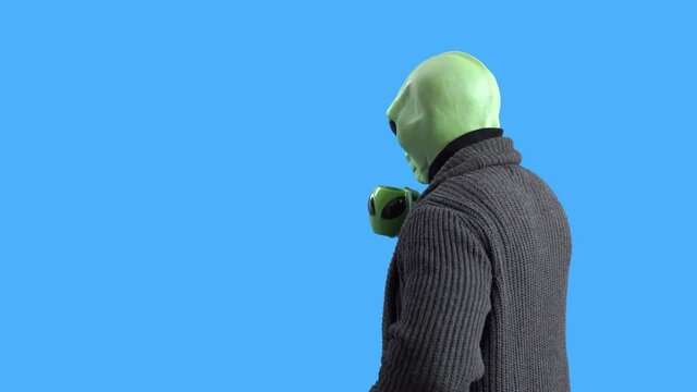 Man in alien costume, warm knitted jacket, with funny themed mug in his hand pretends to wipe the window with sleeve and drink hot beverage against a blue chroma key. Preparation for Halloween party.