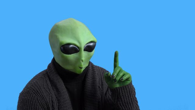 Old Retired Alien In Warm Knitted Jacket Instructively Raises Index Finger In The Air And Nods His Head Against Blue Chromakey Background. Themed Costume For Party.