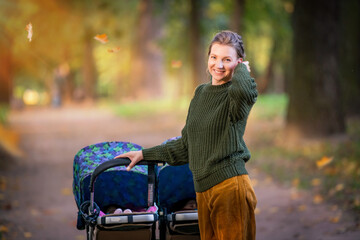 Happy mom are walking in autumn park with a stroller for twins. Full-length portrait.