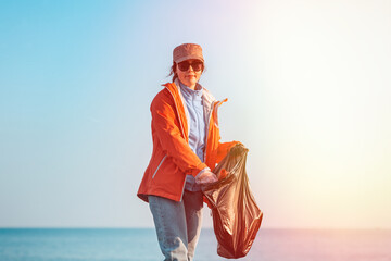 A young Caucasian woman in a jacket collects garbage and puts it in a plastic bag. In the background, the horizon of the sea and sky. Copy space and light