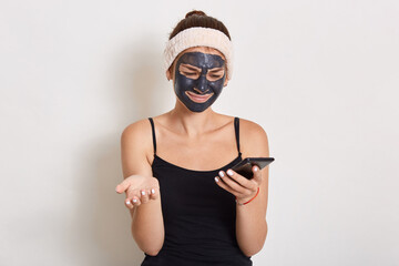 Young lady with black facial cosmetic mask using cellphone, looking directly at device screen, spreading palm, having astonished and puzzled facial expression.