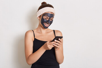 Smiling girl reading message on smart phone with black mask on her face, wearing hair band and...