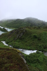Fototapeta na wymiar Berglandschaft mit Wiese und grüner Alm und See und Fluss im Nebel mit Wolken