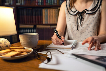 A woman is sitting at a table and writing something in a notebook.On the table is a laptop, cookies and a Cup of tea. Close up.The concept of quarantine, remote work and freelancing