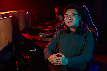 Portrait of Asian young gamer in headphones smiling at camera while sitting on chair and playing computer games