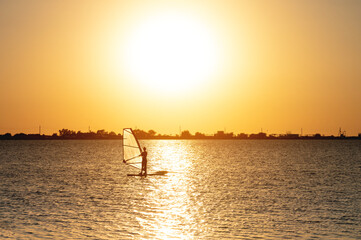 A beginner windsurfer woman stands on a board with a sail on a sunset background. Windsurfing school