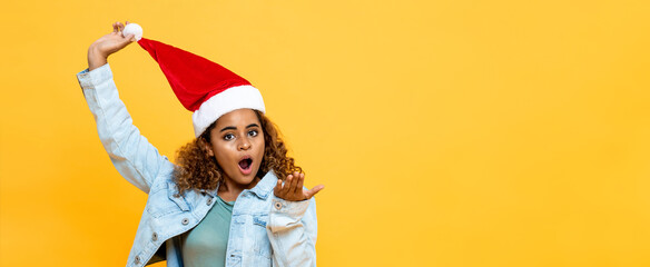Surprised African American woman wearing Christmas hat with open palm gesture on yellow isolated...