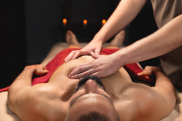 A male massage therapist makes a massage of the pectoral muscles and chest. Sports massage for a muscular client in a dark room by candlelight