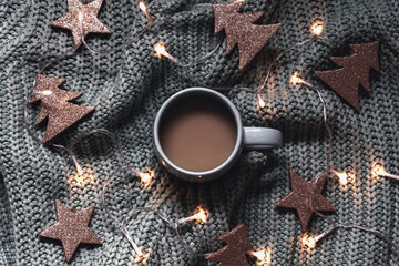 A cup of coffee with christmas lights and toys on grey knitted sweater background. Winter holidays concept. Top view, flat lay.