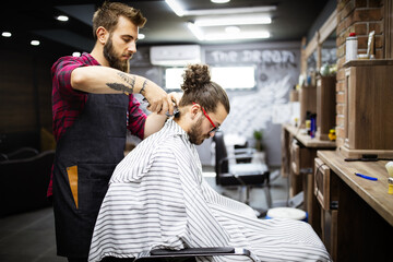 Young bearded man getting haircut by hairdresser with electric razor at barber shop