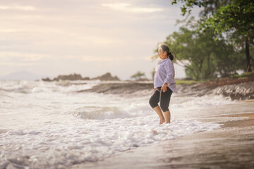 Senior Asia woman walking on the beach with barefoot, Smile with happily while wave in the sea rush to shore.