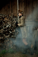 Beautiful sexy brunette girl in a coat and a hat with exquisite makeup on the background of an old wooden building with a stack of firewood and smoke from a fire