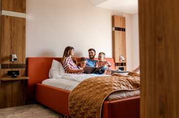 Family of four enjoying together at hotel room, sitting at bed reading hotels offers.