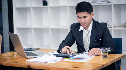 A handsome young Asian businessman working at home with a laptop and calculator documents on the desk according to the concept of a holiday businessman.