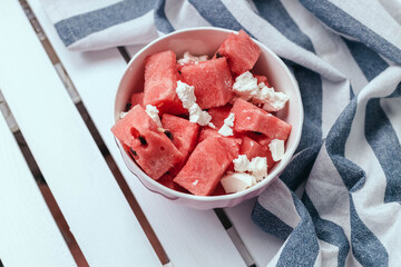 Delicious summer salad with slices of watermelon and feta cheese on white bowl with a striped towel on a wooden white background