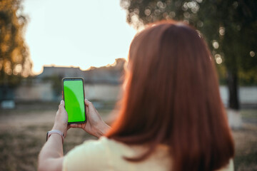 Young girl with her back makes selfie on a smartphone in the park. Mockup blank screen on the phone
