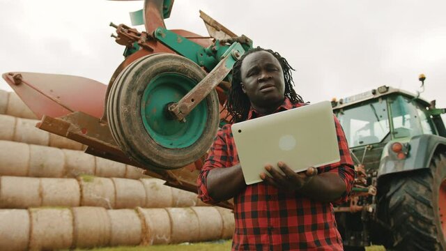 Young african farmer using tablet in front og big green tractor anf haystack. High quality 4k footage