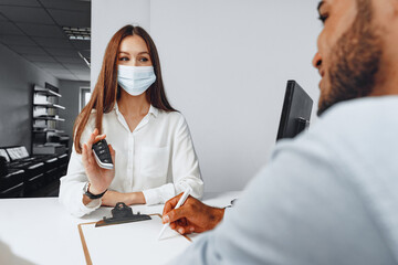 Attractive woman car seller wearing mask gives keys to car buyer