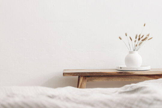 Modern white ceramic vase with dry Lagurus ovatus grass and marble tray on vintage wooden bench, table. Blurred beige linen blanket in front. Scandinavian interior. Empty white wall, copy space.