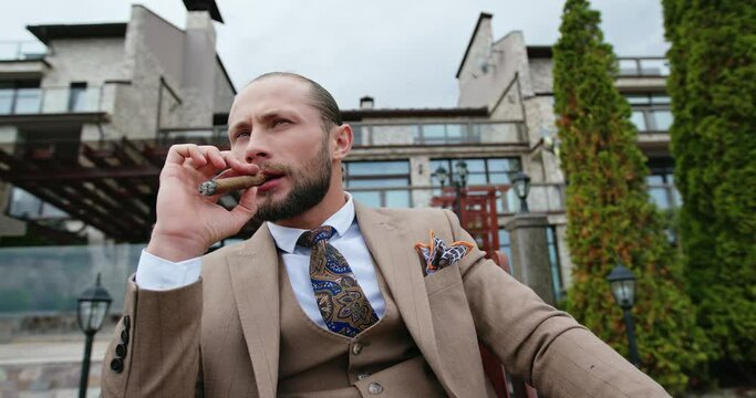 Male businessman dressed in a suit. Serious smoking a cigar in the yard of his house - slow motion