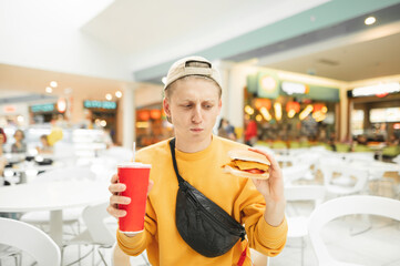 Funny young man stands with a burger and a glass of cola in his hand and looks upset at a fast...