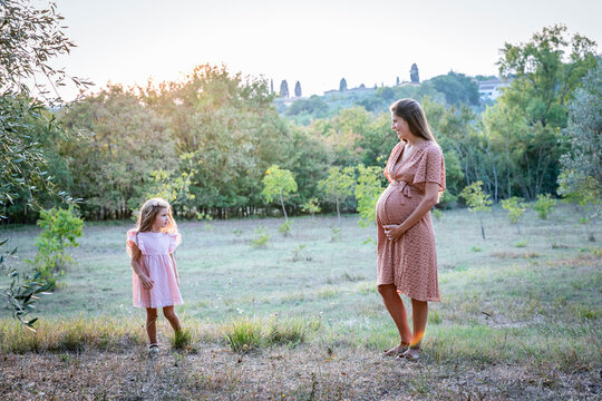 Expecting woman with little daughter standing in green field