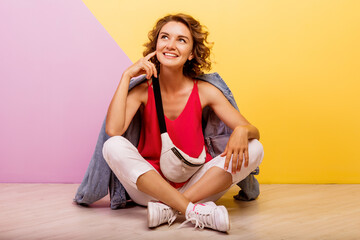 Fototapeta na wymiar Studio image of smiling brunette lovely woman wearing stylish sporty outfit and jeans jacket sitting on the floor. Pink and yellow background.