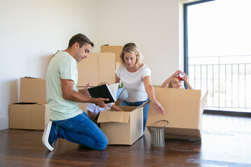 Focused parents and funny kids unpacking things in new apartment, sitting on floor and taking objects from open box. House buying or moving concept