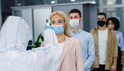 Fototapeta premium Health control at work and new normal for company. Man in protective suit checks temperature with infrared thermometer at multiracial workers in protective masks