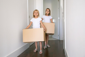 Fototapeta na wymiar Sweet pretty girls moving in new flat, carrying carton boxes in corridor. Front view, full length. Apartment buying concept
