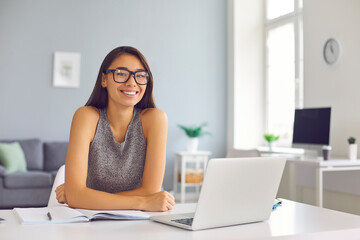 Happy young woman in cool eyeglasses sitting at desk with open laptop and notebook looking at camera