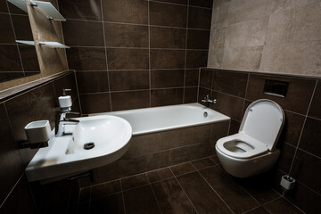 Small bathroom of the city apartment with wash basin, bath, toilet and all furnishing. Paving and tiling have a texture of concrete trowel in a two shades of brown color. The toilet seat is open.