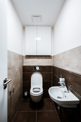 Small toilet of the city apartment with all furnishings, including a small sink, cabinet and ceiling fan. Paving and tiling have texture of concrete trowel in shade of brown. The toilet seat is open.