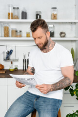 young tattooed man in white t-shirt reading newspaper in kitchen