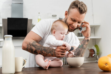 Obraz na płótnie Canvas young father talking on smartphone during breakfast with little son in kitchen