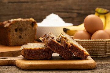 Sliced ​​banana cake is placed on a wooden tray on a wooden table, with a banana, egg and flour as a background.