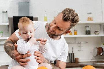 selective focus of baby boy licking spoon near young father in kitchen